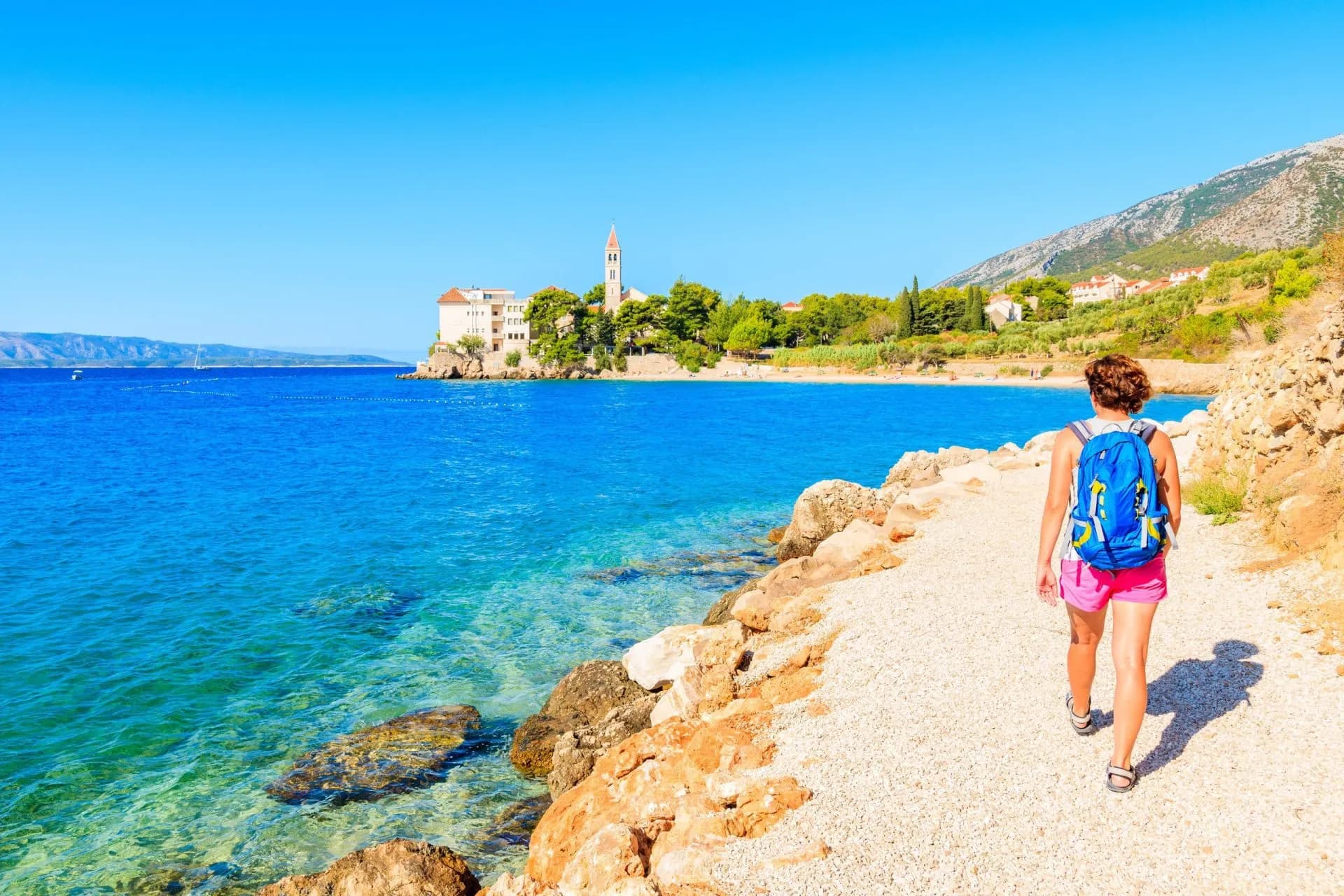 Hiker walking path by turquoise sea toward coastal town with church tower in Croatia